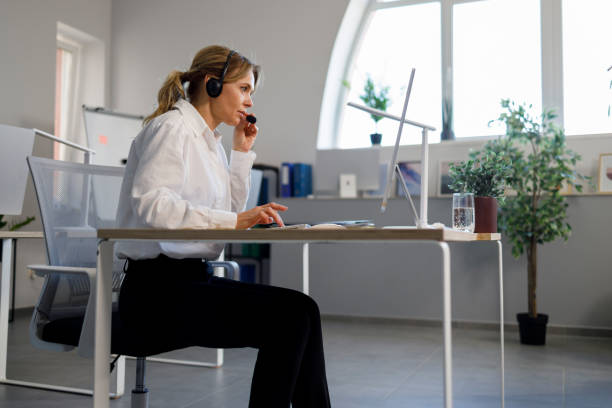 wanita bisnis dewasa melakukan panggilan video, menggunakan headset. tampilan samping operator pusat panggilan yang duduk di tempat kerja di kantor - agen judi online potret stok, foto, & gambar bebas royalti