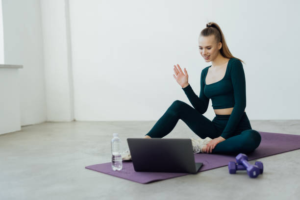 istock Online Exercise Session Marathon, Remote Training. Happy millennial female sitting on floor mat and talking with coach, having videocall with teacher, using pc for virtual workout 1394981434