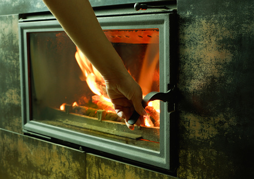 A woman's hand opens the glass door of the fireplace in which the wood is burning. Modern closed fireplace with glass.