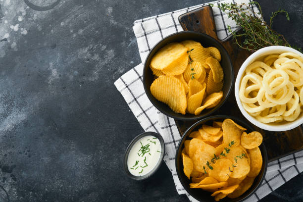 ungesundes essen. schnellimbisse. alle klassischen kartoffelsnacks mit erdnüssen, popcorn- und zwiebelringen und gesalzenen brezeln in schüsseltellern auf schwarzem betonhintergrund. ungesunde nahrung für figur, herz, haut, zähne - onionskin stock-fotos und bilder