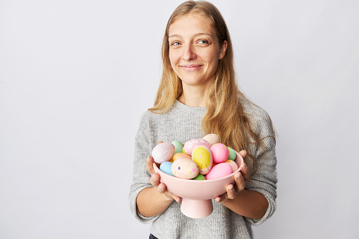 A young woman with blond long hair and blue eyes holds a bowl with colorful Easter eggs in her hands while standing in front of a gray wall
