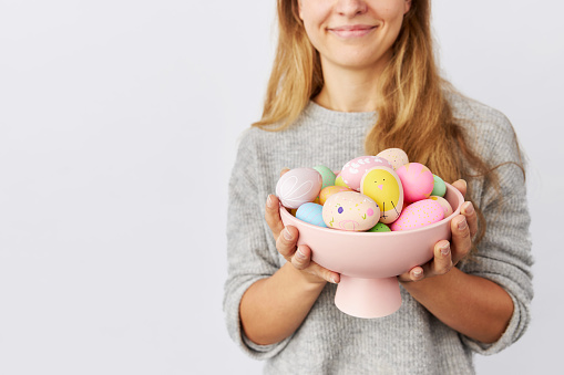 A young woman with blond long hair and blue eyes holds a bowl with colorful Easter eggs in her hands while standing in front of a gray wall