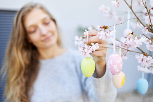 A young woman with blond long hair and blue eyes happily hangs easter eggs on her cherry tree in the garden