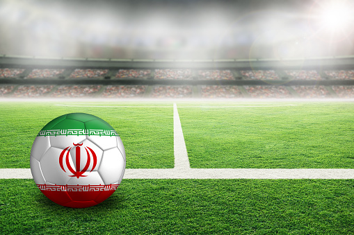 Football in brightly lit outdoor stadium with painted flag of Iran. Focus on foreground and soccer ball with shallow depth of field on background and copy space.