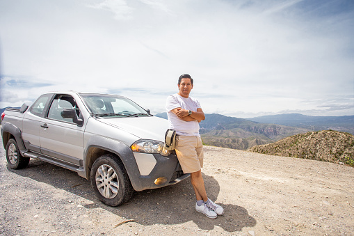 Young adult wearing a white t-shirt, brown shorts and white tennis shoes, leaning on his gray truck on the side of the road and in the background a mountain range, while making a trip alone.