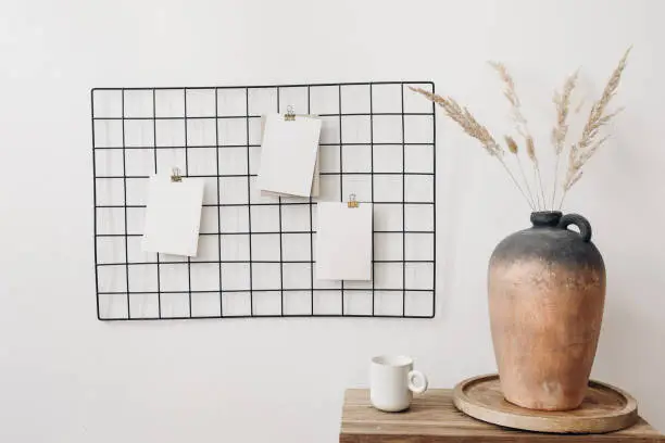 Black metal mesh noticeboard, bulletin board with blank memo cards mockups. Elegant home office interior concept. Vase with dry lagurus grass and cup of coffe on wooden table, white wall background.