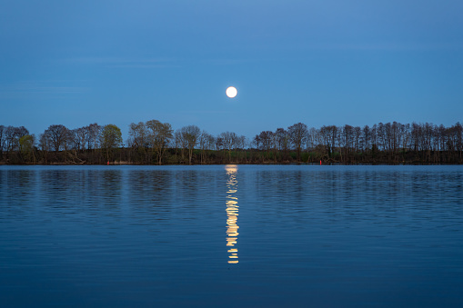 Moonrise over a tranquil lake landscape view. Bright shining moon reflected on the water surface. Idyllic evening in the nature. Night environment with calm waters.