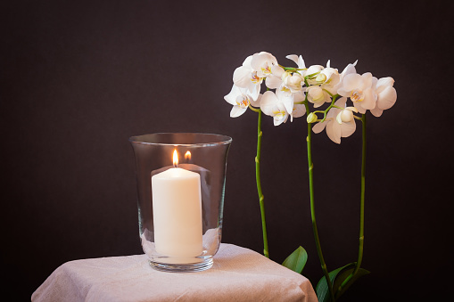 Burning candle in glass vase and white orchid on dark background