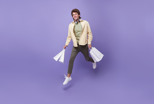 Handsome man jumping fast carry many bags rushing next shop store isolated over purple background.
