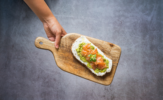Top view anonymous hand holding a delicious cheese, avocado and salmon toast on wooden cutting board