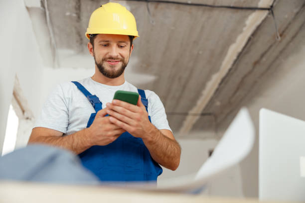 istock Focused repairman in workwear and hardhat smiling while using smartphone, standing indoors during renovation work 1395015800