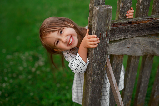 A cheerful funny little girl looks out from behind an old wooden gate in the backyard. Natural expressive emotions of a child. Happy baby. Copy space