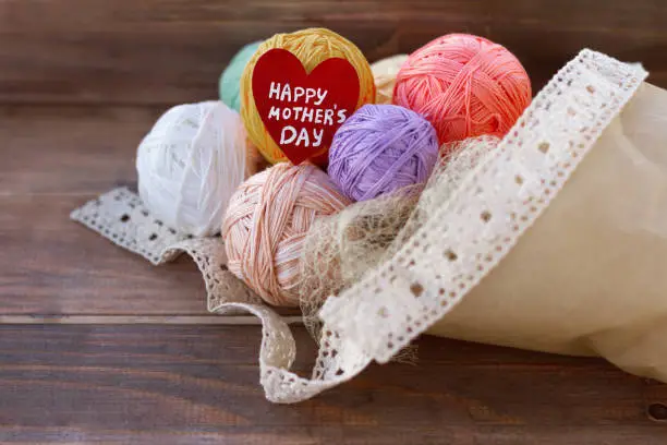 Photo of red wooden heart and happy mother's day lettering, bouquet of multi-colored balls of yarn in craft packaging, cotton ribbon on wooden table