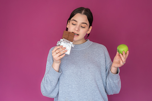 A young woman with apple and chocolate on pink background, food and diet concept.