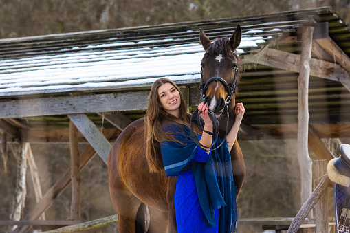 A girl in a blue dress hugs a horse against the backdrop of a snow-covered canopy in the forest