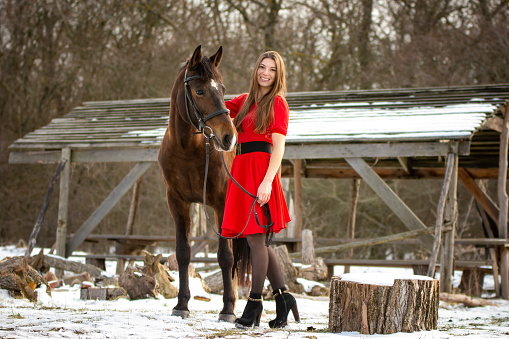 A beautiful girl in a red dress walks with a horse in the rays of the setting sun against the backdrop of old ruins