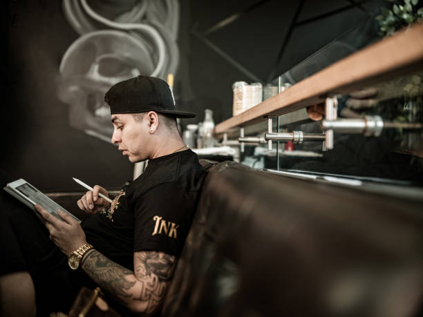 Young male Mexican Tattoo artist sketching on digital tablet. He is wearing t-shirt with his own design logo print and baseball hat. Interior of tattoo parlour during day.