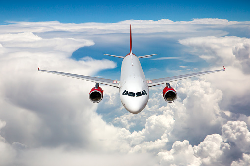White passenger plane with red engines in flight. Aircraft flies high in the blue sky above the clouds. Front view.