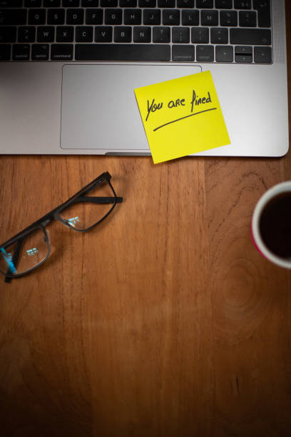 Workspace with a laptop, pen and glasses and a post-it note stuck on the computer keyboard with a message in English: you're fired