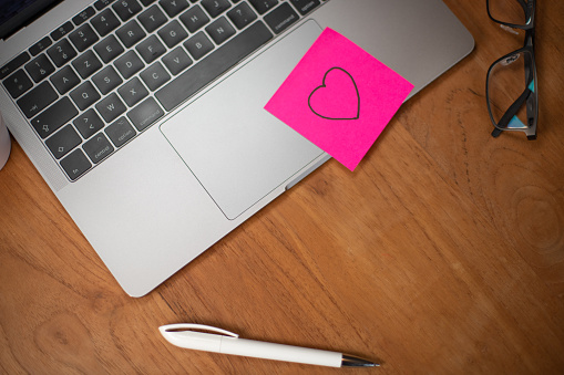 desk, workspace with a laptop. A pink post-it note with a heart drawn on it is stuck on the computer keyboard. Symbol of happiness, well-being at work.