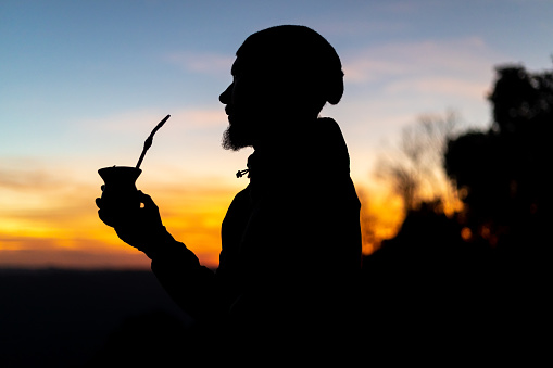 Silhouette of a man with a cap drinking yerba mate, traditional South American drink, at sunset. Twilight sky
