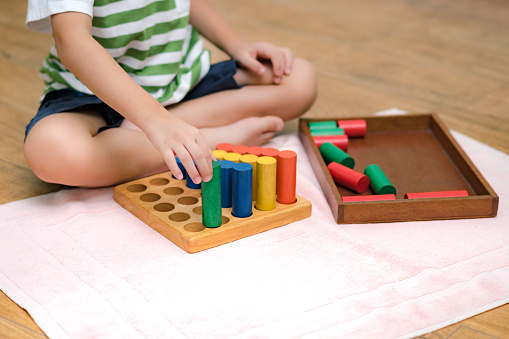 Cute Asian Montessori preschool boy student sitting, learning and playing with colorful wooden cylinder socket in classroom.