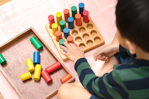 Cute Asian Montessori preschool boy student sitting, learning and playing with colorful wooden cylinder socket in classroom.