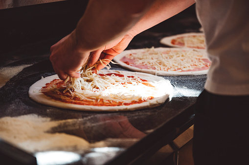 Persons hand adding toppings on pizza, adding freshly grated mozzarella cheese on the pizza lying on black granite counter.