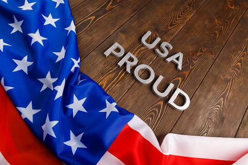 words USA proud laid with silver metal letters on brown wooden surface with flag of United States of America in diagonal view.