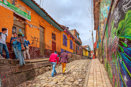 Bogotá, Colombia - July 20, 2016: A few tourists and some local Colombians seen on the broader end of the, colourful, cobblestoned Calle del Embudo which is at the opposite end of the Plaza del Chorro de Quevedo. The Street gets its name from its funnel shape: Embudo translates to Funnel in English. It is shaped like a funnel. It must be remembered that these streets were set up over 450 years ago when people usually travelled on horseback. The Street is well known for its Street Art, many of which feature the legends of the Pre-Colombian era. Photo shot in the late afternoon sunlight; horizontal format.