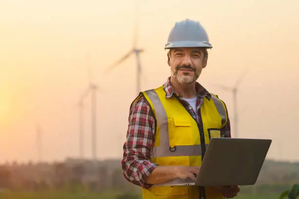 Photo of Portrait of power engineer wearing safety jacket and hardhat with laptop computer working at outdoor field site that have wind turbine at the background.