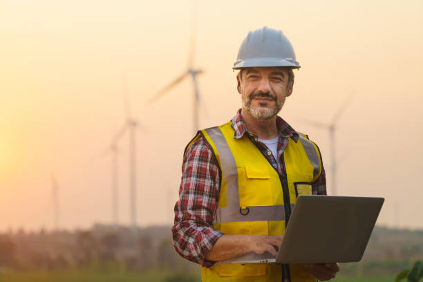 portret inżyniera energetyka w kamizelce ochronnej i kapeluszu z laptopem pracującym na terenie zewnętrznym, który ma turbinę wiatrową w tle. - engineer zdjęcia i obrazy z banku zdjęć