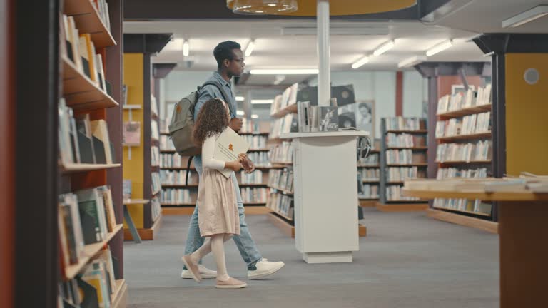 Biracial father and daughter walking through a library together. Diverse family browsing the library.