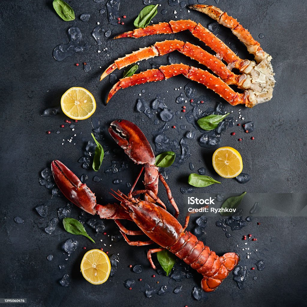 Lobster and crab legs with ice and lemon on dark background top view. Delicatessen crab and lobster seafood on black slate table. Crustacean seafood aesthetics. Luxury food - alive lobster and crab legs. Lobster and crab legs with ice and lemon on dark background top view. Delicatessen crab and lobster seafood on black slate table. Crustacean seafood aesthetics. Luxury food - alive lobster and crab legs Lobster - Seafood Stock Photo