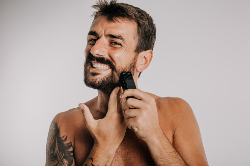 Grooming concept. Young man shaving his beard with a trimmer or electric shaver against the white background.