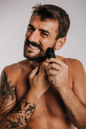 Grooming concept. Young man shaving his beard with a trimmer or electric shaver against the white background.