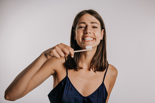 Photo of a cute, happy, woman, a brunet,  holding a toothbrush. Starting to brush her teeth. while standing in a white background studio. Copy space. Dental care