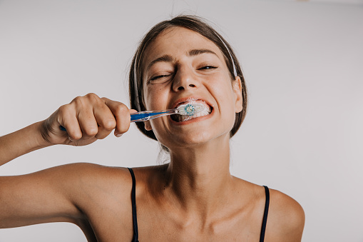 A headshot photo of a cute, happy, young adult woman, a brunet, brushing her teeth with a big morning smile on her face, facing the mirror, in a white background studio. Dental care
