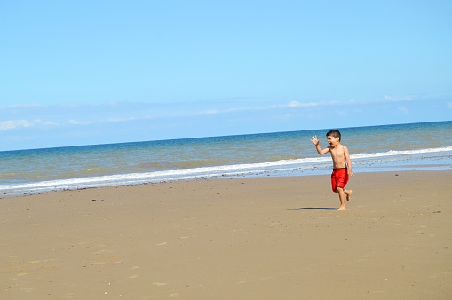 Horizontal photograph of a small child running  on the wet sandy beach. The boy is barefoot and topless. The little boy in red colored swimming trunks with a black band has no shirt, bare bodied, body. There is Copy space for text. There is sea and sky meeting in the horizon. The kid is happy and waving his hand.