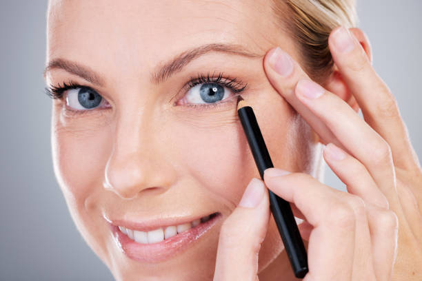 retrato de estudio de una atractiva mujer madura que aplica delineador de ojos sobre un fondo gris - makeup pencils fotografías e imágenes de stock