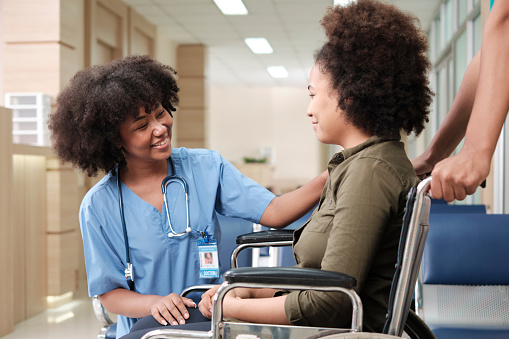 Young African American female doctor with stethoscope in uniform checks injury patient girl in wheelchair at outpatient accident clinic hospital, illness medical clinic examination, healthcare hall.