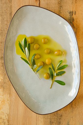 Olives and olive oil.Green olives and olive branch in olive oil in a ceramic gray plate close-up on a natural light wood table.Cold Pressed Organic Natural Oil