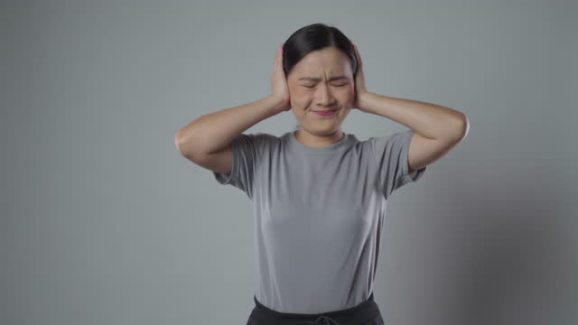 Asian woman bored and annoyed, covering her ears isolated over gray background.
