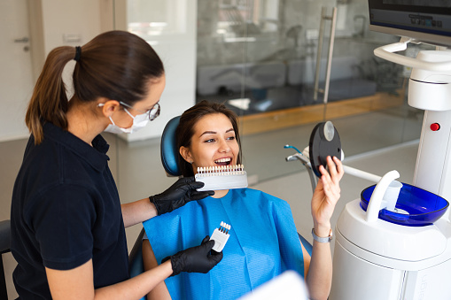 A female dentist wearing a surgical facemask uses a dental tooth shade guide for whitening teeth service to ensure a perfect choice of color for the beautiful young patient. Bleaching, prosthetics, orthopedic dentistry concept.