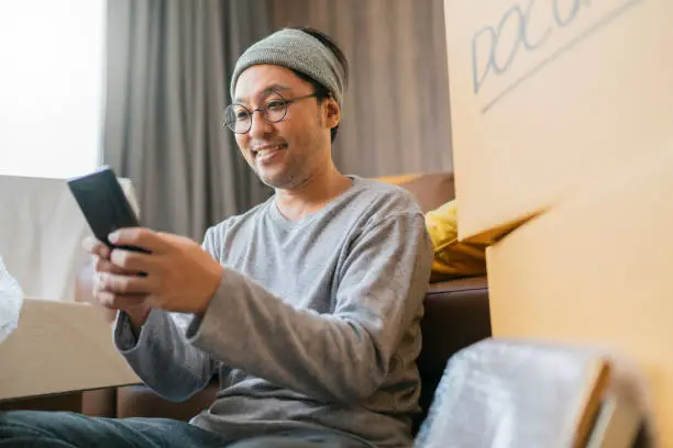 asian mature adult male man is packing and unpacking as he is moving into a new place house. He is using his smartphone to talk and chat text to friends and family.home moving ideas concept