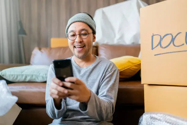 asian mature adult male man is packing and unpacking as he is moving into a new place house. He is using his smartphone to talk and chat text to friends and family.home moving ideas concept