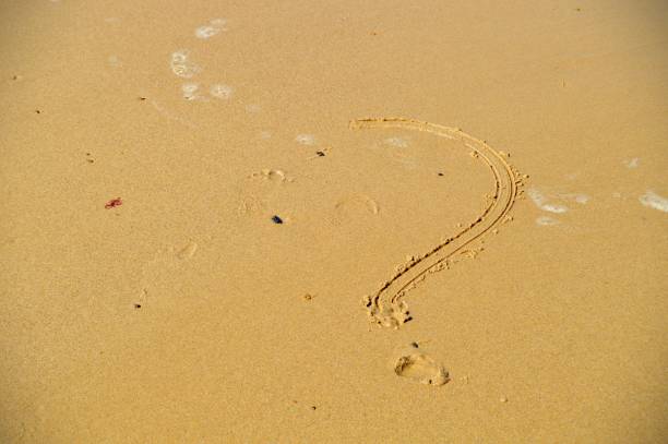 Question mark hand written on wet sand on a beach on a sunny day. Horzontal image. The question mark is handwritten in double line. The dot below the question mark is like a small pit. A couple of black and brown pebbles are also on sand.