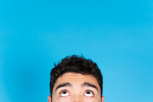 Half portrait of hispanic teenager boy looking up to copy space isolated on blue background.