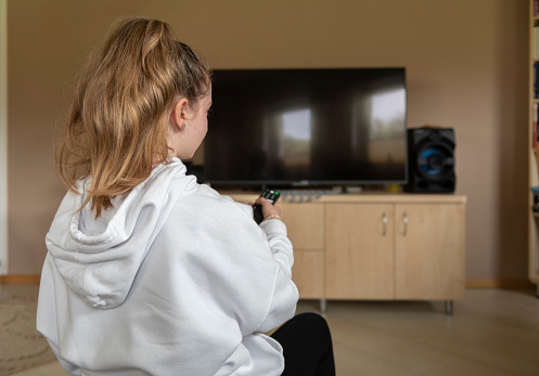 Back view of teenage girl sitting on sofa at home turning on TV with remote control
