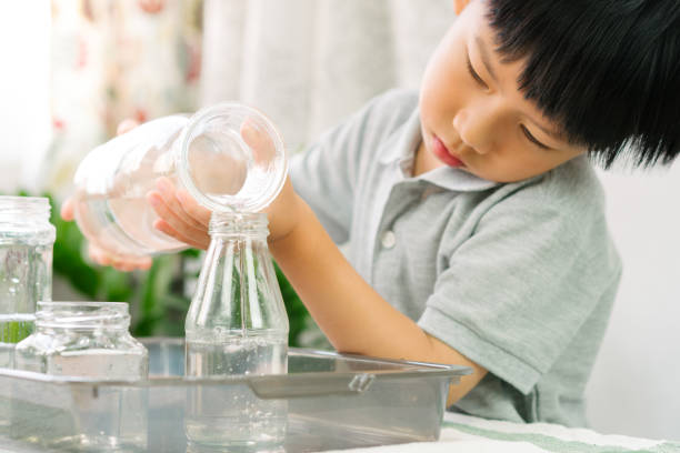 Cute Asian little Montessori boy pouring water into glass water bottles. Cute Asian little Montessori boy pouring water into glass water bottle with concentration on wooden table at home brain jar stock pictures, royalty-free photos & images